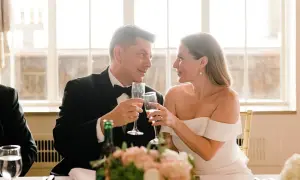 Para młoda wznosi toast. W tle okna, a na stole bukiet kwiatów. Idealny moment na piosenki na wesele.