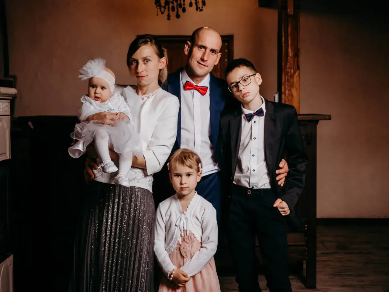 Rodzina świętuje chrzest święty. Mama trzyma niemowlę w białej sukience, obok stoi starsza córka, a rodzice i starszy syn w garniturach.