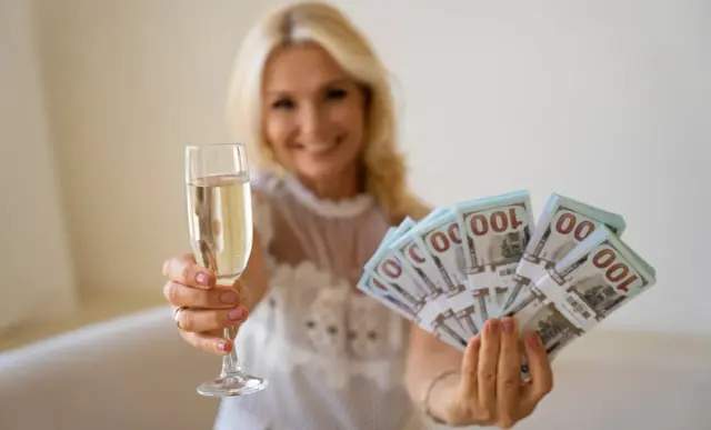 Uśmiechnięta blondynka trzyma kieliszek szampana i stos banknotów 100 dolarowych. Ile się daje na 18? Tyle, by świętować!